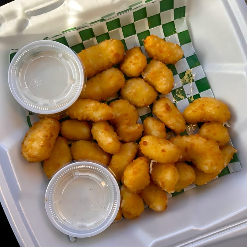 Golden fried cheese curds with two dipping sauces in a white takeout container with green checkered paper.